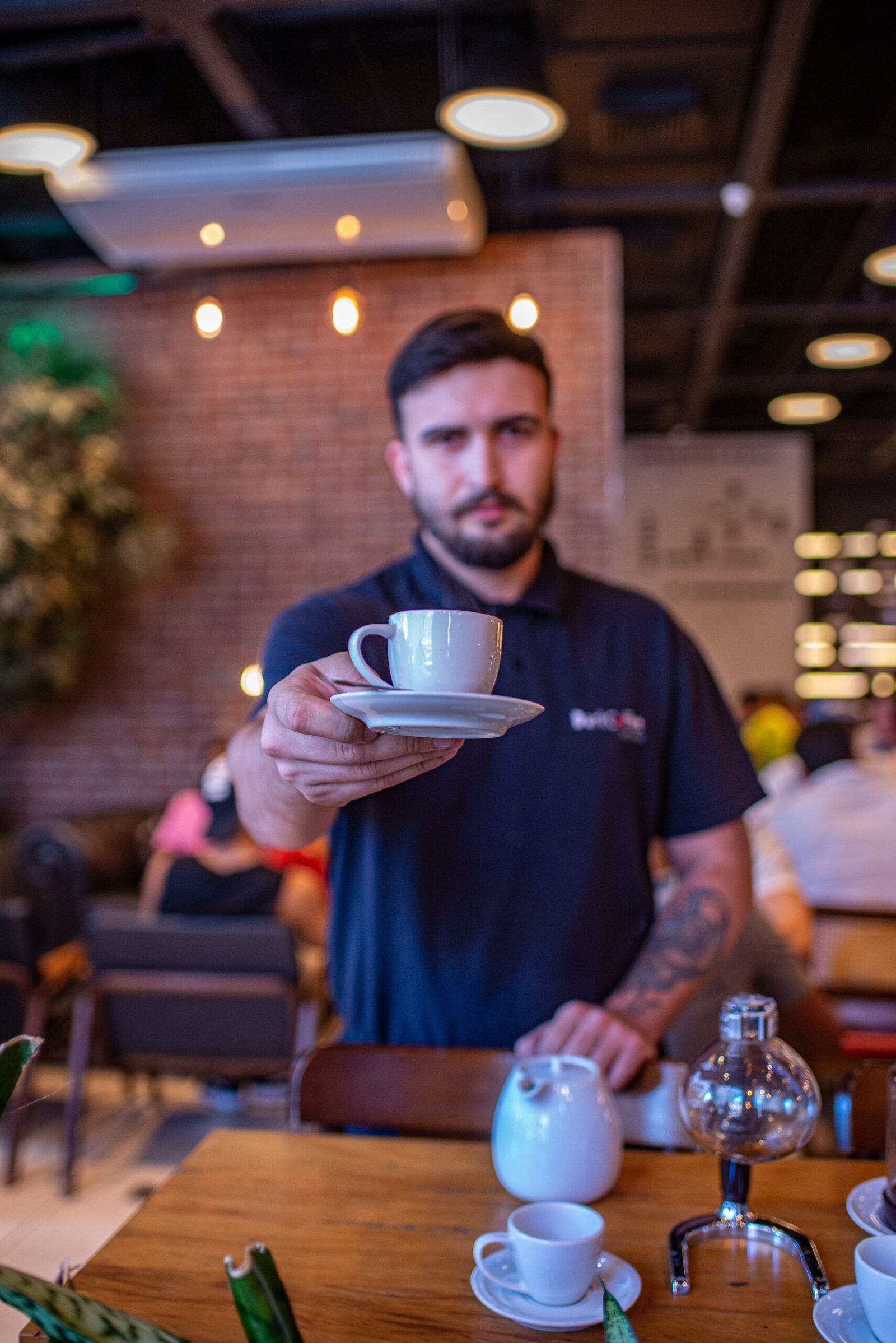 Home A barista in a modern café extends a cup of coffee towards the camera, creating a welcoming atmosphere.